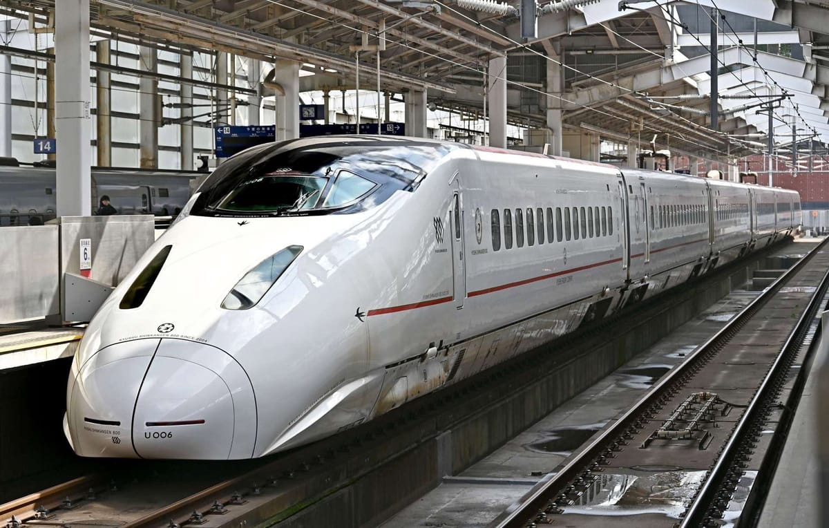 九州新幹線 800 系列車停靠在車站月台，白色車身配紅色線條，上方是車站鋼架屋頂
